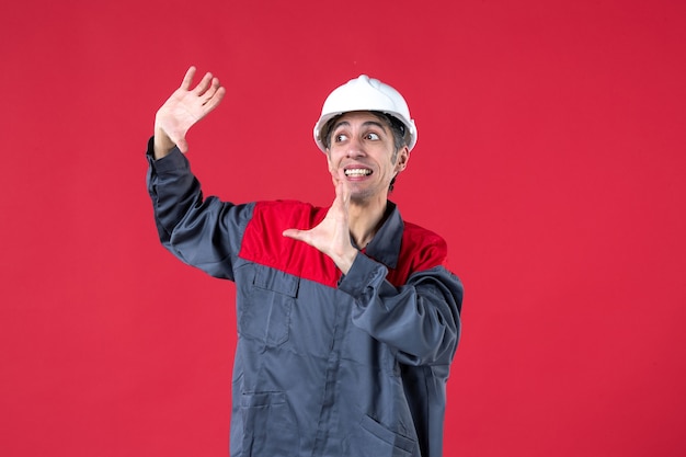 Side view of surprised young worker in uniform with hard hat on isolated red wall