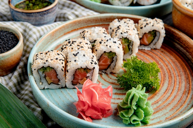 Side view of sushi rolls with tuna salmon and avocado covered with sesame on a plate with wasabi and ginger