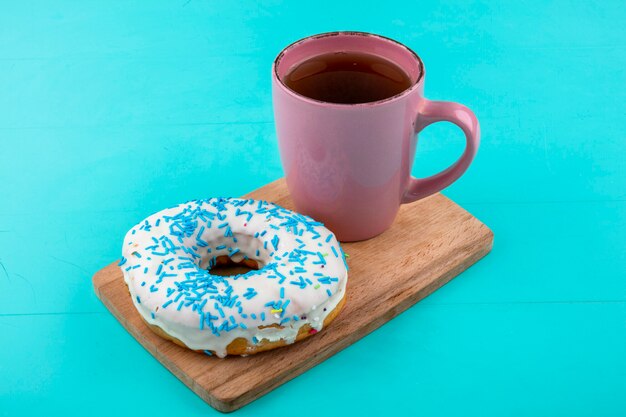 Side view of sweet donut with a cup of tea on a cutting board on a turquoise surface