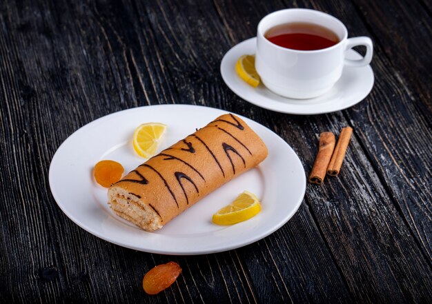 Side view of swiss roll with apricot jam on a white plate served with a cup of tea on rustic