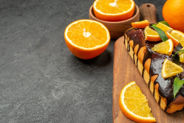 Вид сбоку на вкусные торты целиком и нарезанные апельсины на разделочной доске на черном столе