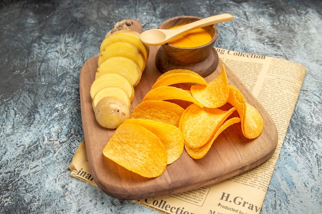 Side view of tasty homemade chips cut potato slices on wooden cutting board on newspaper on gray table