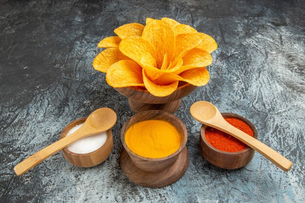 Side view of tasty potato chips decorated like flower shaped different spices with spoons on them on gray table