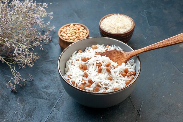 Side view of tasty rice meal with beans in a white small pot and its ingredients wooden spoon on blue background