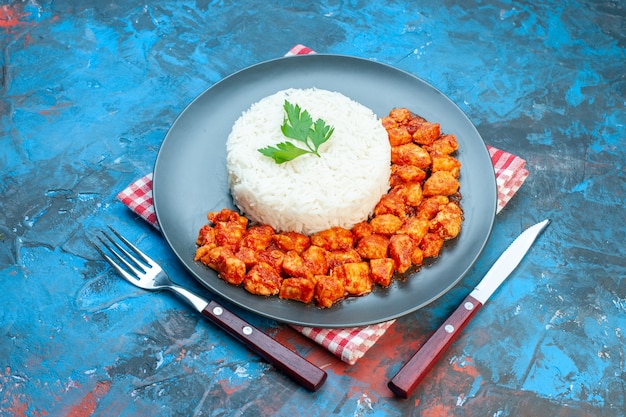 Side view of tasty rice meal with green and tomato chicken on red stripped towel next to cutlery set on blue table