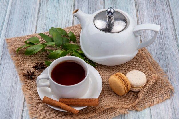 Side view of teapot with a cup of tea cinnamon and macarons on a beige napkin on a gray surface