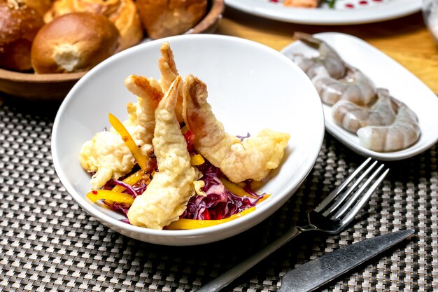 Side view of tempura shrimps with chopped red cabbage and peppers in a white bowl on the table