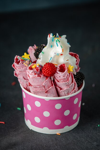Вид сбоку тайского мороженого ролл под взбитыми сливками украшен ягодами и красочными брызгает в картонной миске на черной стене