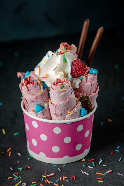 Side view of thai ice cream roll under whipped cream decorated with berries and colorful sprinkles in a cardboard bowl on black wall