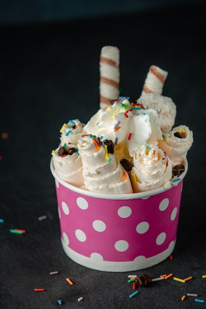 Side view of thai ice cream roll under whipped cream decorated with colorful sprinkles in a cardboard bowl on black wall