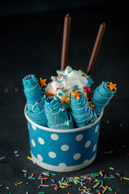 Side view of thai ice cream roll under whipped cream decorated with colorful sprinkles in a cardboard bowl on black wall