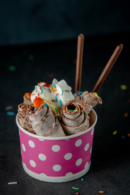 Side view of thai ice cream roll under whipped cream decorated with colorful sprinkles in a cardboard bowl on black wall