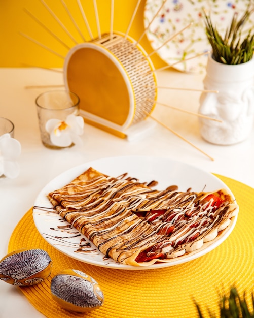 Side view of thin pancake with sliced strawberries and bananas covered with chocolate sauce on white plate