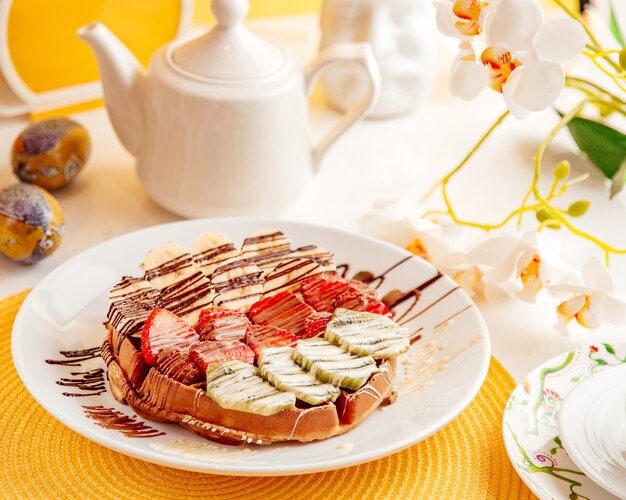 Side view of thin pancake with strawberries bananas and kiwi covered with chocolate sauce on white plate