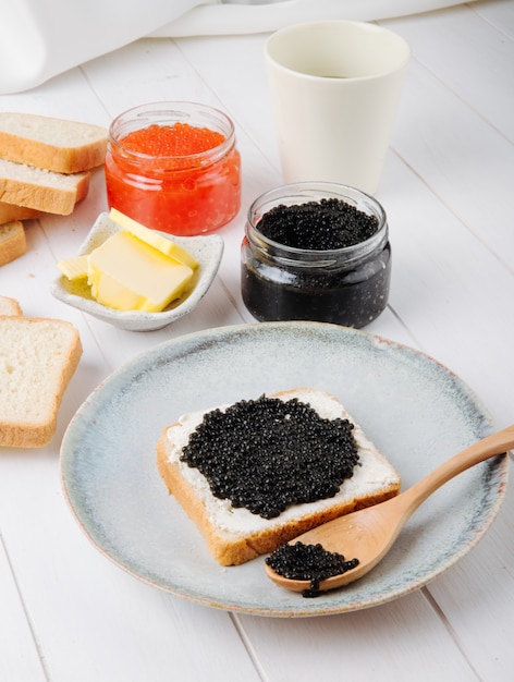 Side view toast with black caviar on a plate with a spoon and butter with a can of black and red caviar