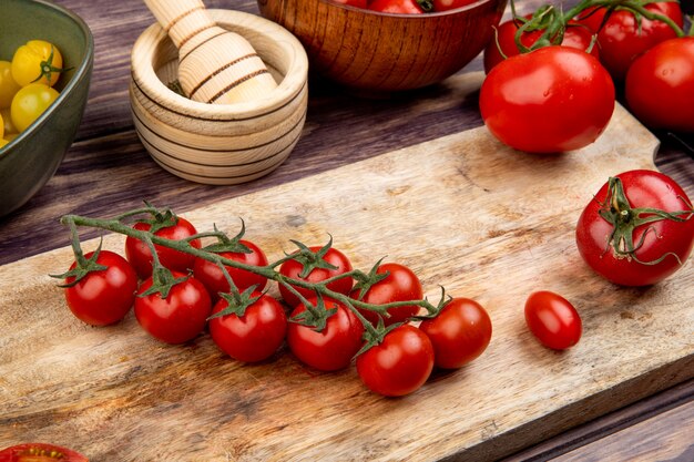 Side view of tomatoes on cutting board with other ones garlic crusher on wooden table