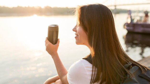 Side view of traveling woman admiring the river while holding thermos