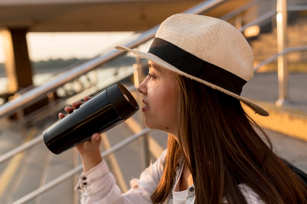 魔法瓶から飲んで帽子を持つ旅行女性の側面図