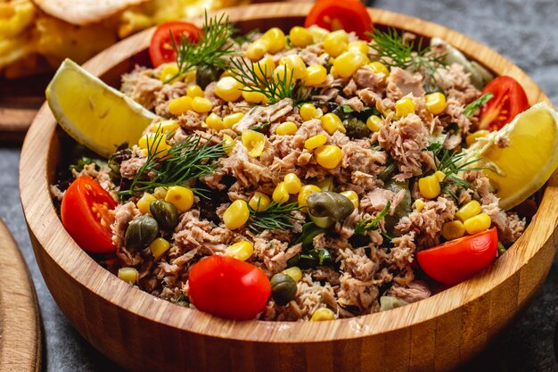 Side view tuna salad with sweet corn pickled capers dill tomato and slice of lemon in a wooden bowl