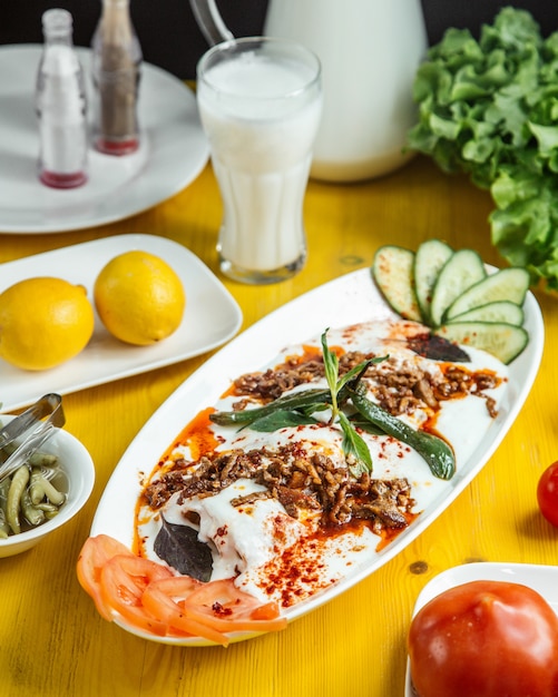 Side view of turkish iskender kebab served with sour yogurt sliced cucumbers and tomatoes on white plate