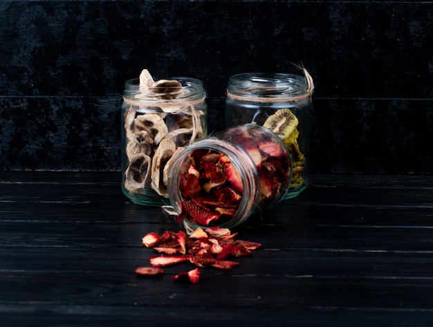 Free photo side view of various dried fruit slices in glass jars strawberry banana and kiwi on black background