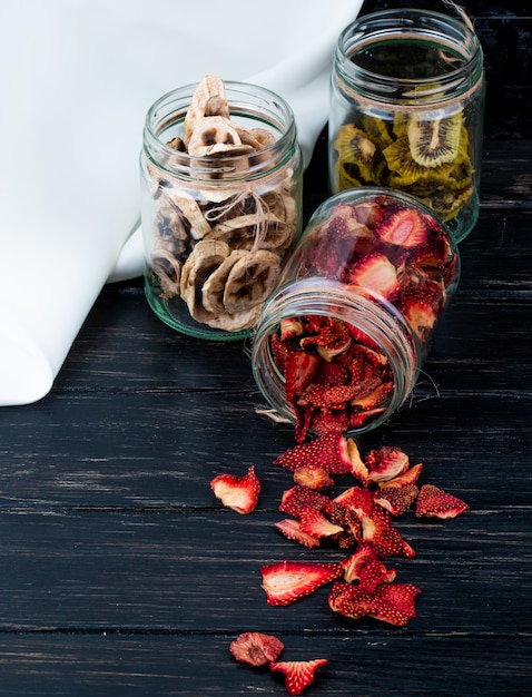 Side view of various dried fruit slices in glass jars strawberry banana and kiwi on black background