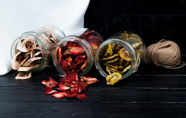 Side view of various dried fruit slices scattered from glass jars on black background