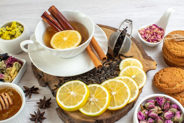 Вид сбоку различных трав и черного чая с медом в белой чашке и лимоном на деревянном подносе, печенье на белом фоне