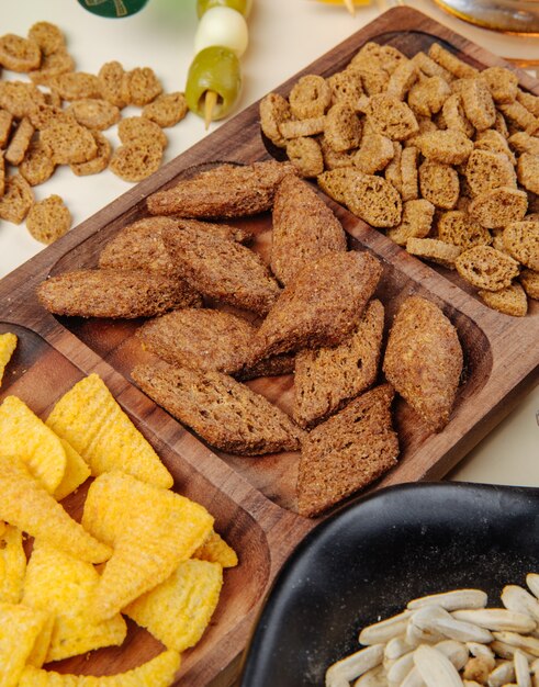 Side view of various salty beer snacks bread crackers corn cones on a wood platter and sunflowers seeds and pickled olives on white