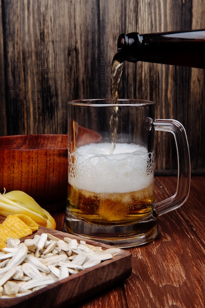 Foto gratuita vista laterale di vari spuntini salati della birra su un vassoio di legno e birra di versamento in una tazza su legno rustico