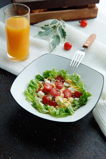 Side view of vegetable salad with tomato egg lettuce in plate with orange juice and leaves with fork on cloth on black background