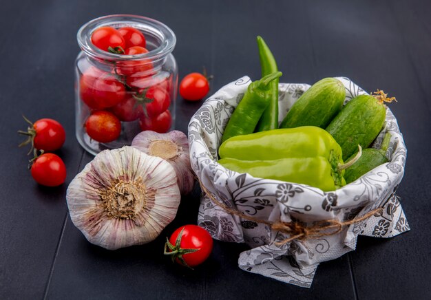 Side view of vegetables as basket of pepper and cucumber and jar of tomato with garlic on black