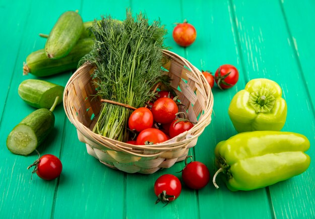 Side view of vegetables as bunch of dill and tomatoes in basket with cut cucumbers peppers and tomatoes on green