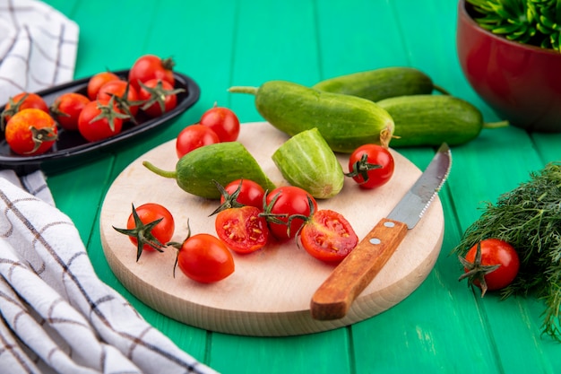 Вид сбоку овощей в виде огурца и помидора с ножом на разделочной доске и пучком укропа и клетчатой ткани на зеленом