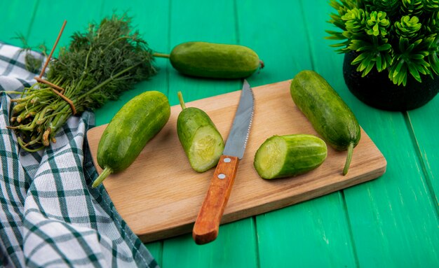 Side view of vegetables as cut and whole cucumbers with knife on cutting board and bunch of dill on plaid cloth and green