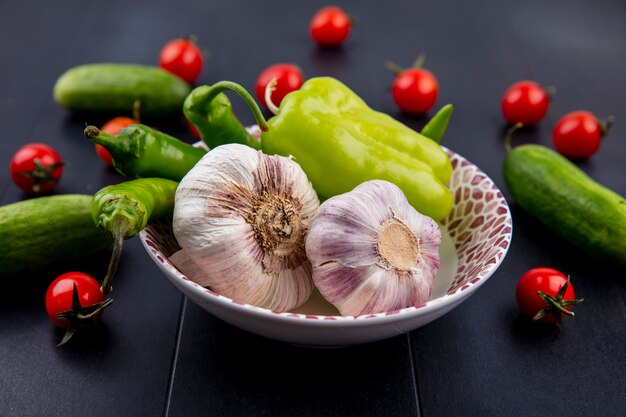 Vista laterale di verdure come aglio pepe nel piatto con pomodori e cetrioli su fondo nero