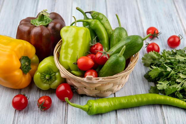 Side view of vegetables as pepper and cucumber in basket with coriander and tomatoes around on wood