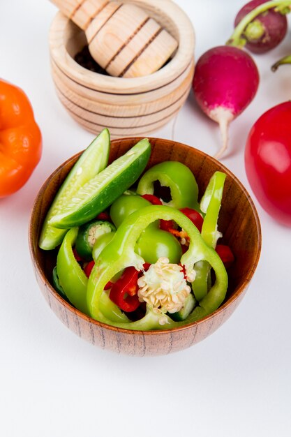Side view of vegetables as sliced peppers and cucumber with radish and tomato with black pepper in garlic crusher on white table