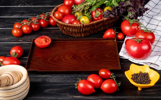 Side view of vegetables as tomato green mint leaves basil in basket and cut tomato in tray black pepper garlic crusher on wooden table