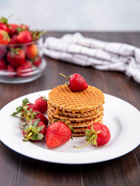 Side view of waffle biscuits and strawberries in plate and in bowl with plaid cloth on wood