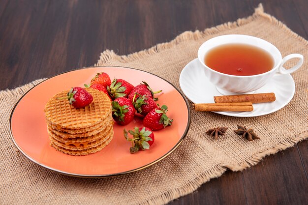 Side view of waffle biscuits and strawberries in plate and cup of tea with cinnamon on saucer on sackcloth and wood