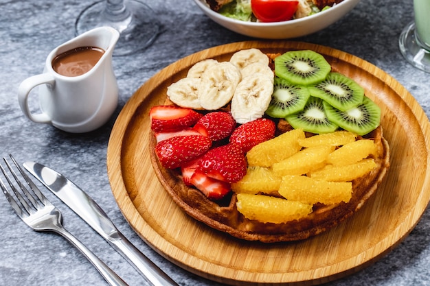 Foto gratuita waffle di vista laterale con l'arancia e il cioccolato della fragola della banana del kiwi sulla tavola