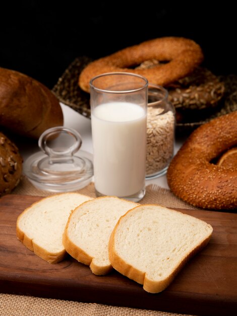 まな板の上の白パンのスライスと黒の背景にパンとオート麦フレークと荒布を着た牛乳のガラスの側面図