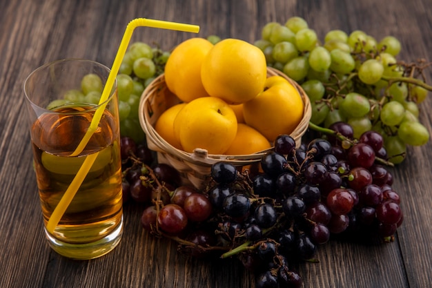 Side view of white grape juice in glass with fruits as nectacots in basket with grapes on wooden background