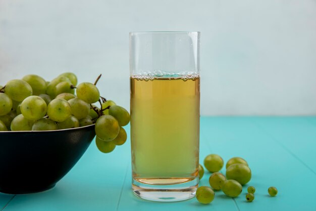 Side view of white grape juice in glass with grape in bowl and on blue surface and white background