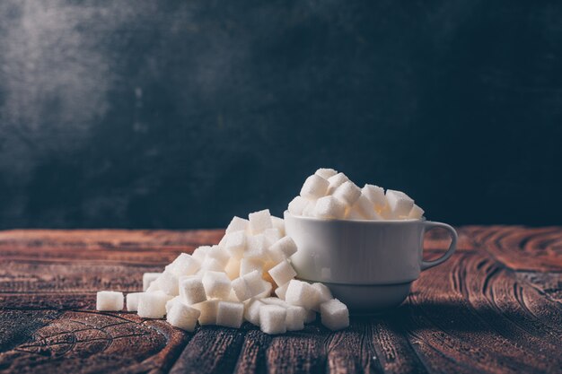 Side view white sugar cubes in a cup on dark and wooden table. horizontal