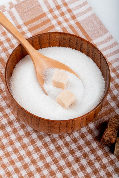 Side view of white sugar in a wooden bowl with a spoon and lump sugar on plaid tablecloth