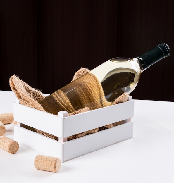 Il vino bianco di vista laterale dentro imbottiglia la cassa di legno e la borsa di tela sulla tavola bianca e sull'orizzontale di superficie marrone