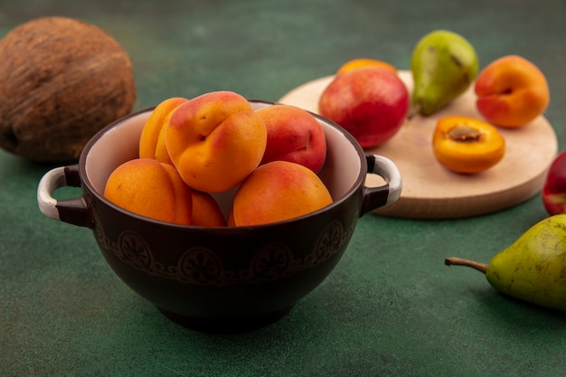 Side view of whole apricots in bowl and half cut one with peach pear on cutting board and coconut on green background