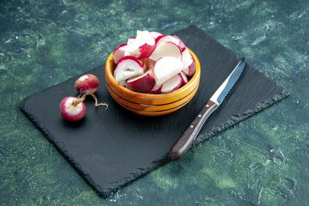 Side view of whole and chopped fresh radishes in bowl on dark color cutting board on green black mixed colors background with free space